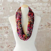 The Veinte Cowl Pattern by Kirsten Hipsky, made with Malabrigo Caracol yarn, is displayed on a white mannequin against a brick wall. It features vibrant pink, orange, black, and white colors with an exquisite texture perfect for any season's style.