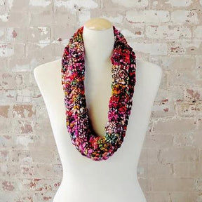 The Veinte Cowl, designed by Kirsten Hipsky, is elegantly showcased on a white mannequin against a textured brick background. Crafted using Malabrigo Caracol with a distinctive knitting pattern, this vibrant multicolored infinity scarf features luxurious superwash merino in shades of pink, red, orange, and black.