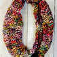 A vibrant and colorful infinity scarf with a thick, chunky texture has been hand-knit from superwash merino. This creation showcases eye-catching hues like pink, purple, orange, and green, beautifully highlighted against a rustic wooden background. Inspired by the Veinte Cowl Pattern by Kirsten Hipsky.