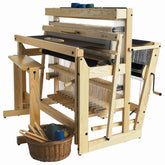 A Louët Delta Loom, complete with a bench, sits ready for weaving. This wooden floor loom from Louët Inc. boasts several spools of thread mounted on it, along with levers, heddles, beams, and an additional pair of Louët Delta Loom Extra Treadles for complex treadling. Beside the loom, a wicker basket filled with additional threads and a few weaving tools completes the setup.