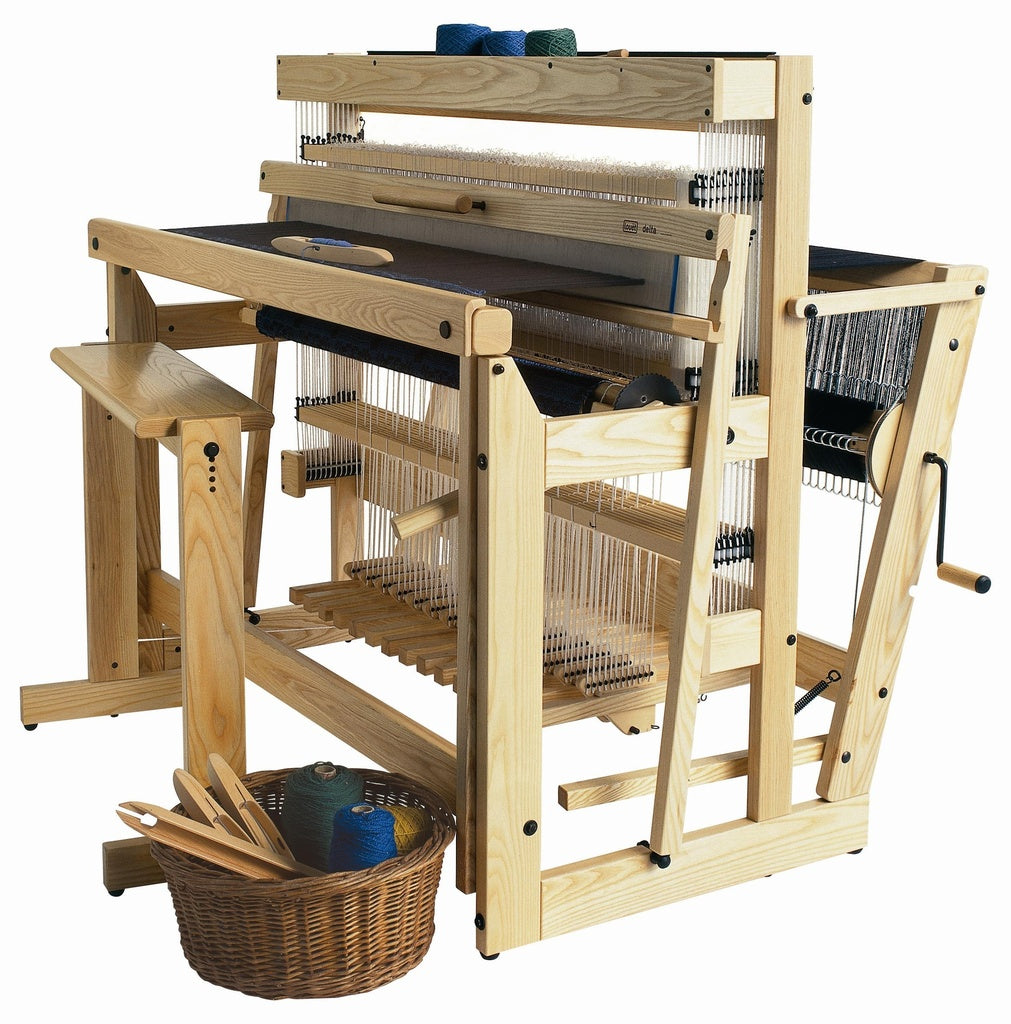 A Louët Delta Loom, complete with a bench, sits ready for weaving. This wooden floor loom from Louët Inc. boasts several spools of thread mounted on it, along with levers, heddles, beams, and an additional pair of Louët Delta Loom Extra Treadles for complex treadling. Beside the loom, a wicker basket filled with additional threads and a few weaving tools completes the setup.