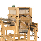 The Louët Megado Treadle Height Adjusting Kit by Louët Inc. is set up, ready for use. A bobbin with green thread is placed on top of the wooden weaving loom, which features a complex arrangement of heddles, shuttles, and beams. With the treadle height adjustment in place, the well-lit apparatus highlights its detailed construction.