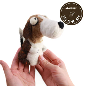 A person holds a felted dog figurine resembling a beagle with brown and white fur. The dog features exaggerated aspects, including large eyes and nose. In the upper right corner, a circular "Woolbuddy" logo is visible, hinting at the "Woolbuddy Needle Felting Kits" used to create this adorable piece.