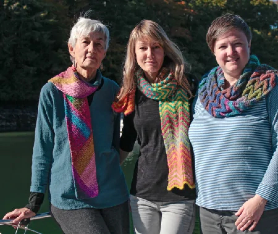 Three people stand close together outdoors, each wearing a colorful knitted scarf. The person on the left has short gray hair and is wearing a blue top. The person in the center has long blonde hair and a black top, and the person on the right has short brown hair and a blue top.