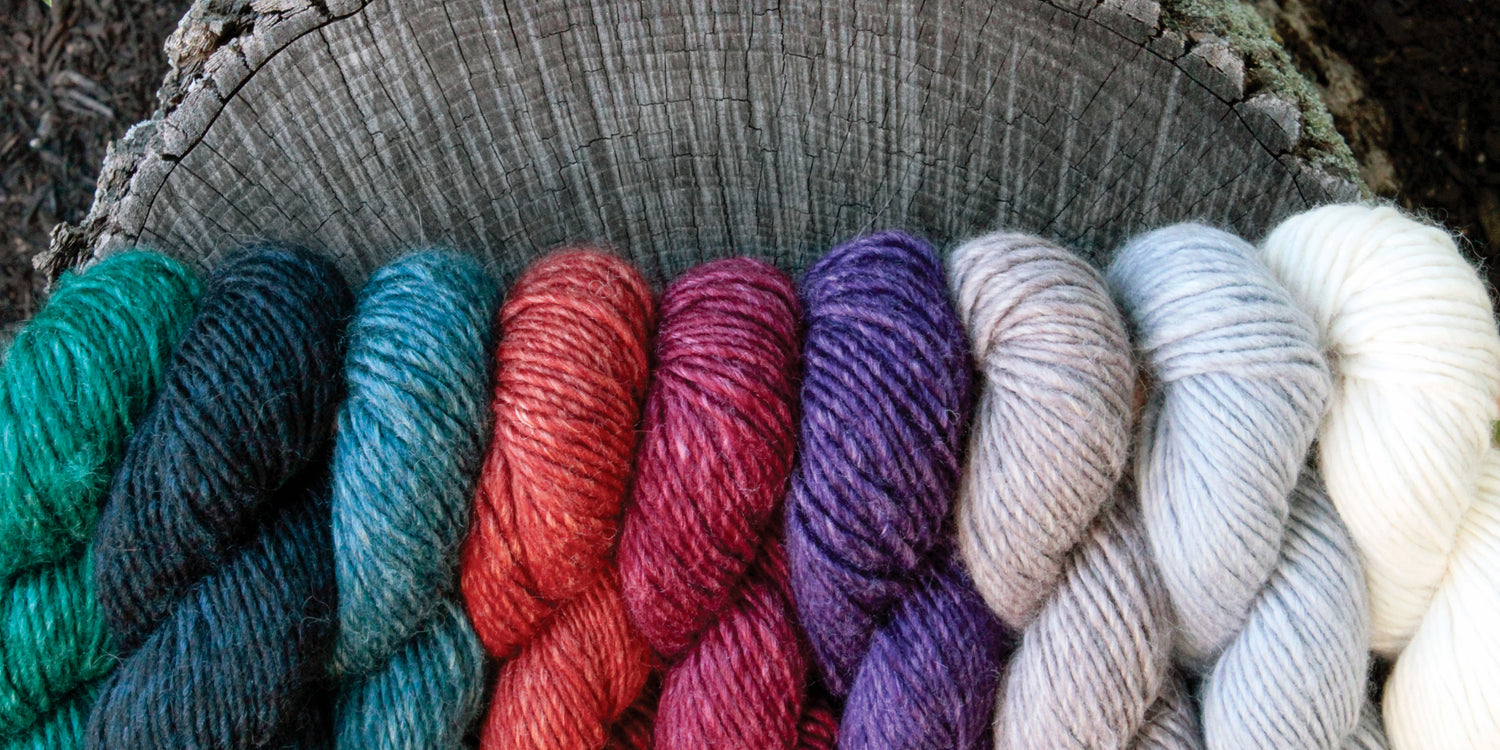 A row of colorful yarn skeins displayed on a tree stump in a close-up view. The yarn colors include various shades of green, blue, red, purple, gray, and white. Each skein is neatly twisted and aligned next to each other.