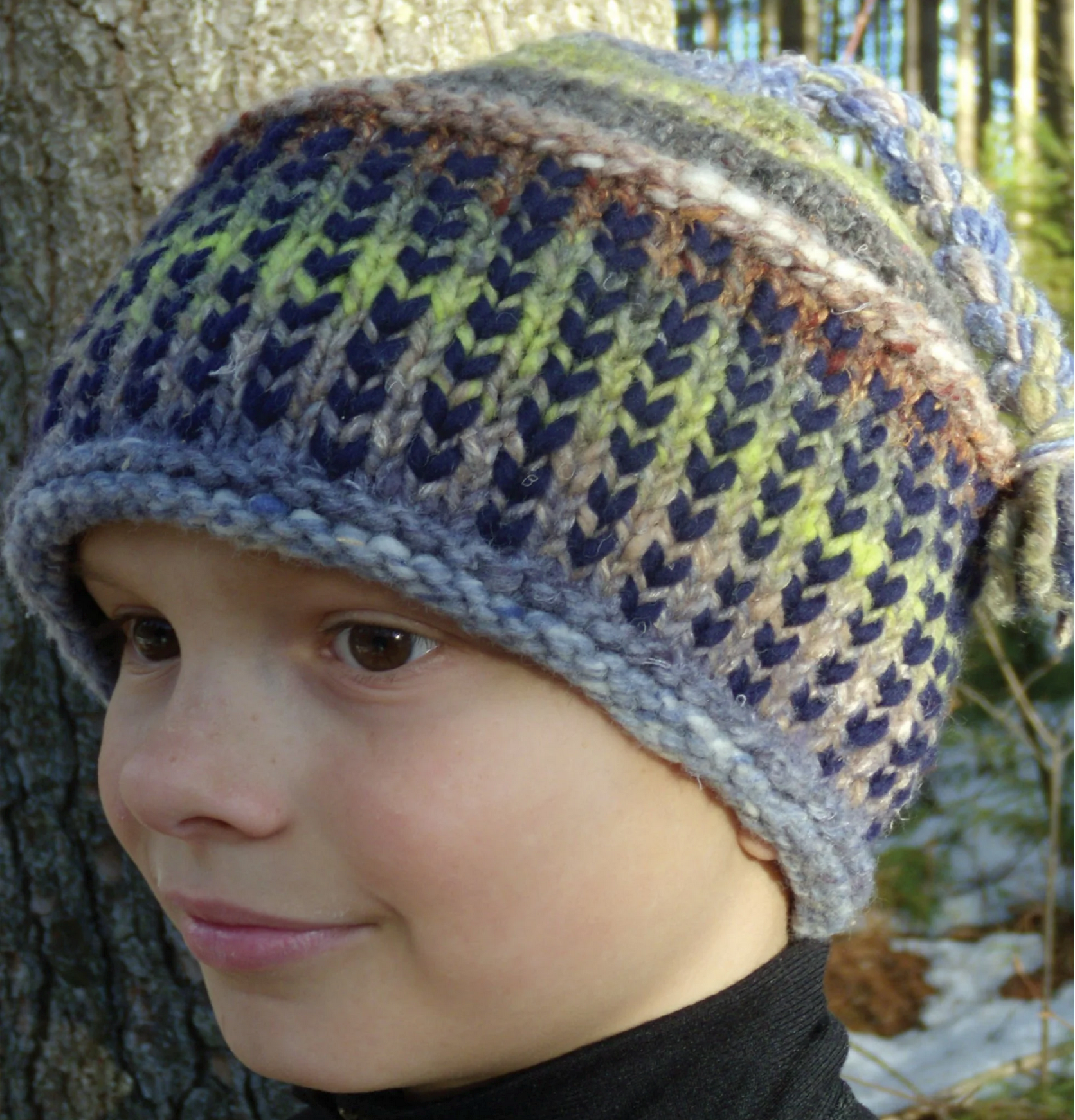 A child wearing a vibrant, knitted wool hat designed with Malabrigo Yarn is gazing off to the side in an outdoor setting. The hat, made from Malabrigo Rasta's super bulky yarn, showcases a pattern of diagonal stitches in blue, green, and brown tones.