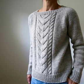 A person stands against a plain background wearing the Avalanche Pullover Sweater, designed by Heidi Kirrmaier, in gray aran yarn with an intricate cable pattern running down the front. The sweater is paired with blue jeans.