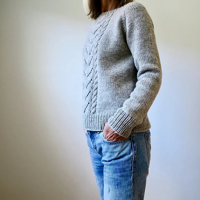 An individual models the Avalanche Pullover Sweater Pattern by Heidi Kirrmaier in gray, paired with blue jeans, set against a simple light background. Their hands are tucked into their pockets, partially obscuring their face.