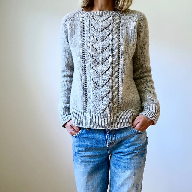 Dressed in the Avalanche Pullover Sweater Pattern by Heidi Kirrmaier, an individual stands with their hands in the pockets of their faded blue jeans against a plain background, head not visible.