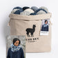 A beige drawstring bag with the "Blue Sky Fibers" logo holds blue and gray yarn balls. The inset shows a person wearing an Aurora Infinity Cowl, exuding woolly warmth. A product tag suggests knitting instructions are included in the Blue Sky Fibers Aurora Infinity Cowl Knitting Kit.