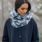 A person in a dark blue coat and thick blue-and-white plaid scarf gazes downward, surrounded by soft, muted trees, evoking the cozy warmth of a Blue Sky Fibers Aurora Infinity Cowl Kit.