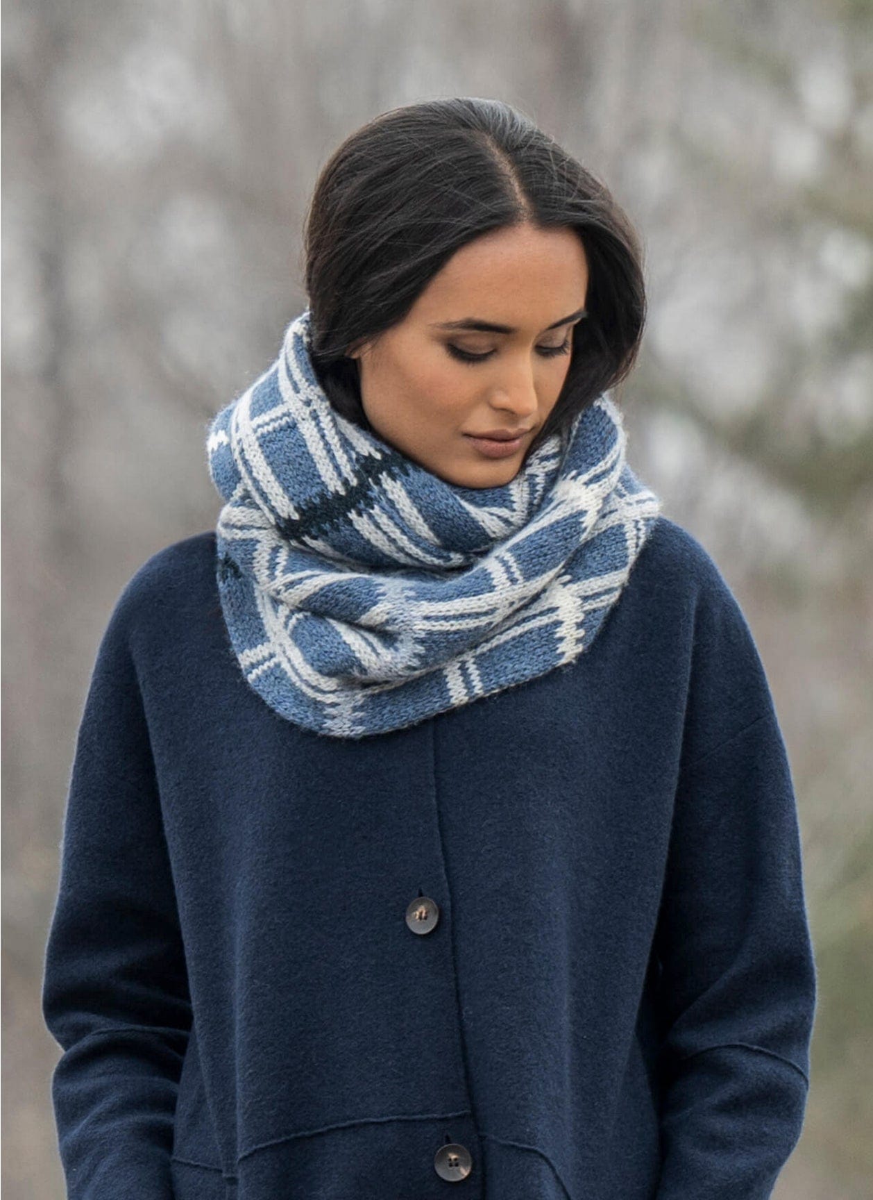 A person in a dark blue coat and thick blue-and-white plaid scarf gazes downward, surrounded by soft, muted trees, evoking the cozy warmth of a Blue Sky Fibers Aurora Infinity Cowl Kit.