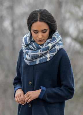 A person wearing a dark blue coat and an Aurora Infinity Cowl from the Blue Sky Fibers Knitting Kit gazes downward. The background is a blurred outdoor setting, possibly in a park or wooded area.