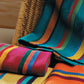 A woven chair is adorned with brightly colored, striped blankets in vibrant green, red, orange, and yellow hues from the "Best of Handwoven Yarn Series: A Dozen Projects in 8/2 Cotton (Printed Version)" by Long Thread Media. Additional folded blankets and patterned napkins rest on the chair's seat and nearby surface.