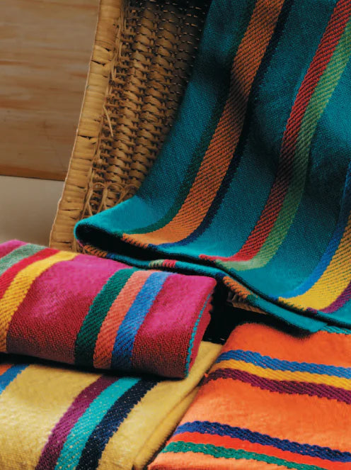 A woven chair is adorned with brightly colored, striped blankets in vibrant green, red, orange, and yellow hues from the "Best of Handwoven Yarn Series: A Dozen Projects in 8/2 Cotton (Printed Version)" by Long Thread Media. Additional folded blankets and patterned napkins rest on the chair's seat and nearby surface.