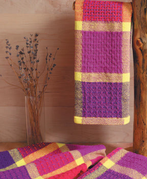 A towel in vibrant checkered patterns, crafted from 8/2 unmercerized cotton in purple, pink, and yellow hues, hangs elegantly from a wooden rod. Below it, additional towels with identical patterns are artfully draped. A vase filled with dried lavender complements the rustic wooden backdrop, highlighting the craftsmanship showcased in Long Thread Media's "Best of Handwoven Yarn Series: A Dozen Projects in 8/2 Cotton" eBook (Printed version).