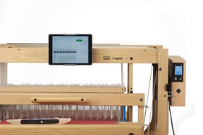 The Louët Megado Floor Loom, produced by Louët Inc., features a wooden frame with a digital interface display panel and an attached tablet, tailored for automated weaving. The loom beautifully combines traditional craftsmanship with modern technology through its visible threads and mechanical components, showcasing intricate weaving patterns.