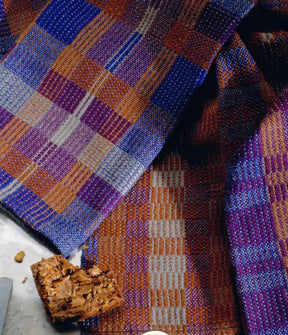 A close-up of a woven textile from the "Best of Handwoven Yarn Series: A Dozen Projects in 8/2 Cotton (Printed Version)" by Long Thread Media, showcasing a vibrant, geometric pattern in shades of blue, purple, brown, and orange. Two pieces of brownies are perfectly positioned on the cloth—one atop the fabric and another partially visible from the side—reminiscent of patterned napkins or placemats and napkin sets.