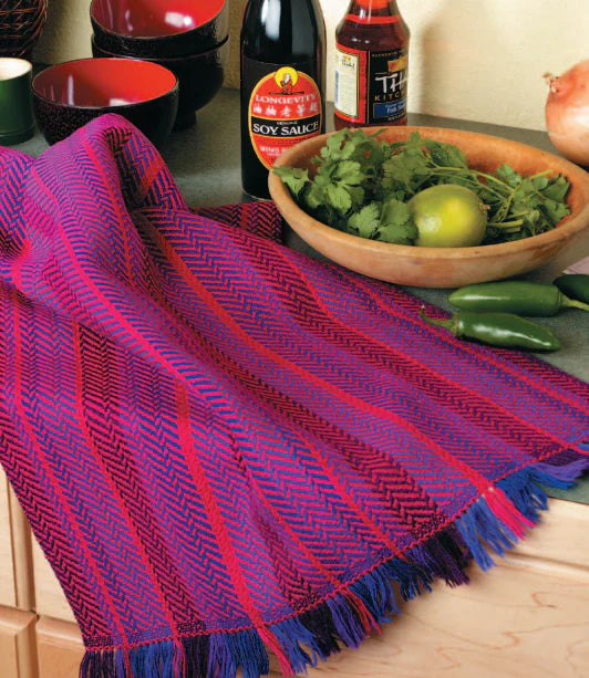 A kitchen counter showcases the vibrant "Best of Handwoven Yarn Series: A Dozen Projects in 10/2 Pearl Cotton" tablecloth by Long Thread Media, complemented by a soy sauce bottle and a wooden bowl containing cilantro, a lime, and a red onion. Nearby, green chilies add to the colorful scene, while assorted bottles and bowls enhance the inviting background.