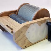 The Majacraft Fusion Engine Drum Carder - 72 TPI, from Majacraft Co., features a wooden frame and a metal handle. The device includes two cylindrical drums with metal teeth and is equipped with a transparent plastic cover on the side. It is designed for preparing wool or other fibers for spinning.