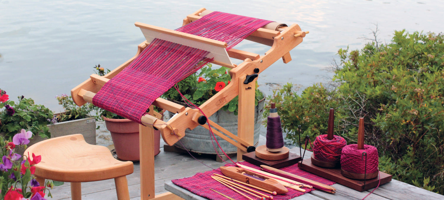 A wooden loom with a magenta and dark blue woven fabric in progress, set on a wooden deck by the water. Nearby, yarn, shuttle sticks, and other weaving tools are arranged. Surrounding the setup are potted plants and greenery.