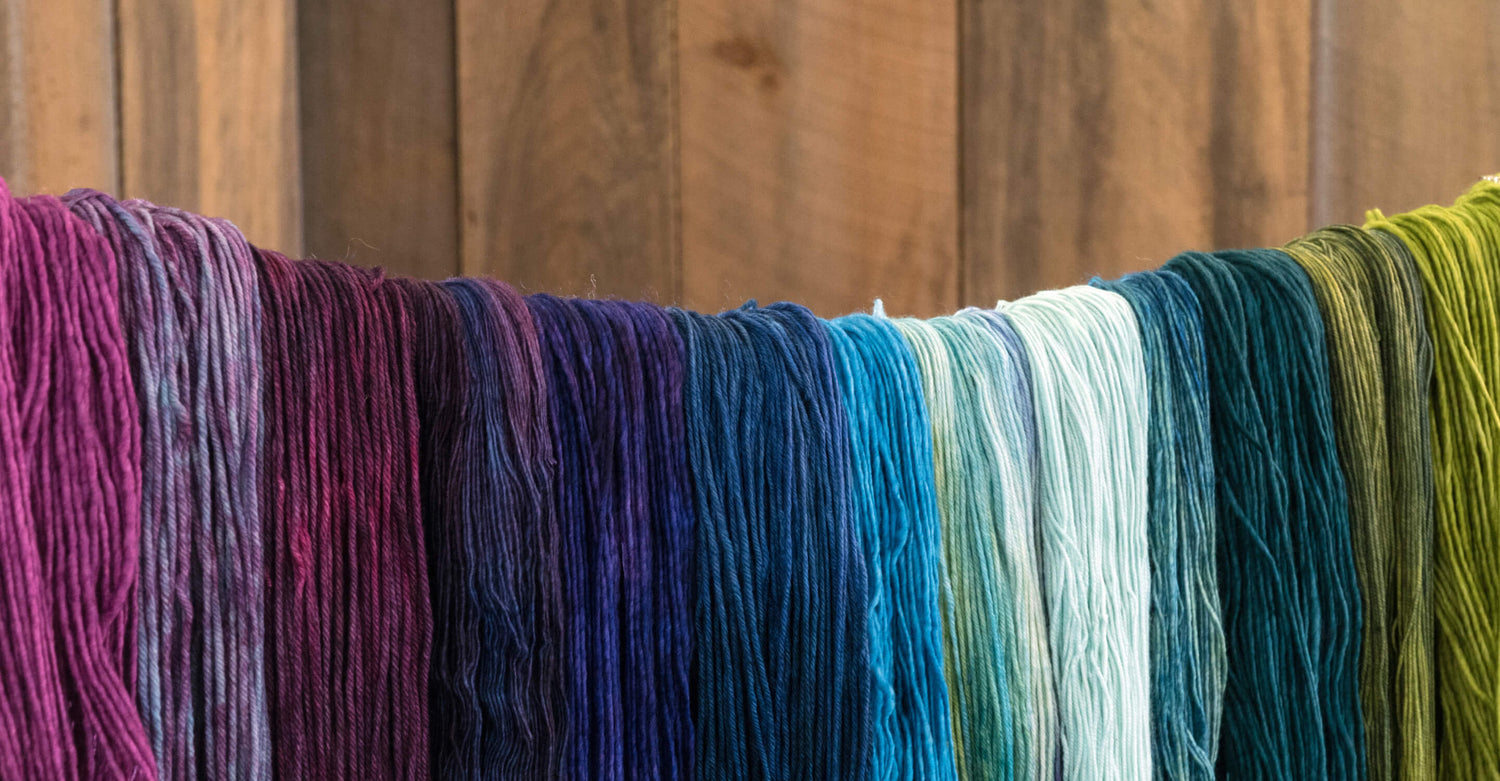 A series of yarns in a spectrum of colors, ranging from deep purples and blues to light blues, greens, and olive tones, are hanging in front of a wooden background. The yarns are draped side by side, showcasing the gradient of hues.