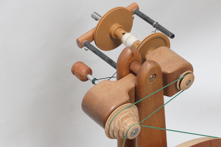 Close-up of a Majacraft Suzie High-Speed Head from Majacraft Co., showcasing the spindle and bobbin mechanisms. The wheel has a natural finish, with fine fiber and green string threaded through parts of the structure. The background is plain and light-colored, emphasizing the details of the spinning wheel.