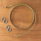 A Leclerc Looms Replacement Flat Steel Brake Circle, with looped ends, sits coiled on a wooden surface next to two metallic S-hooks.