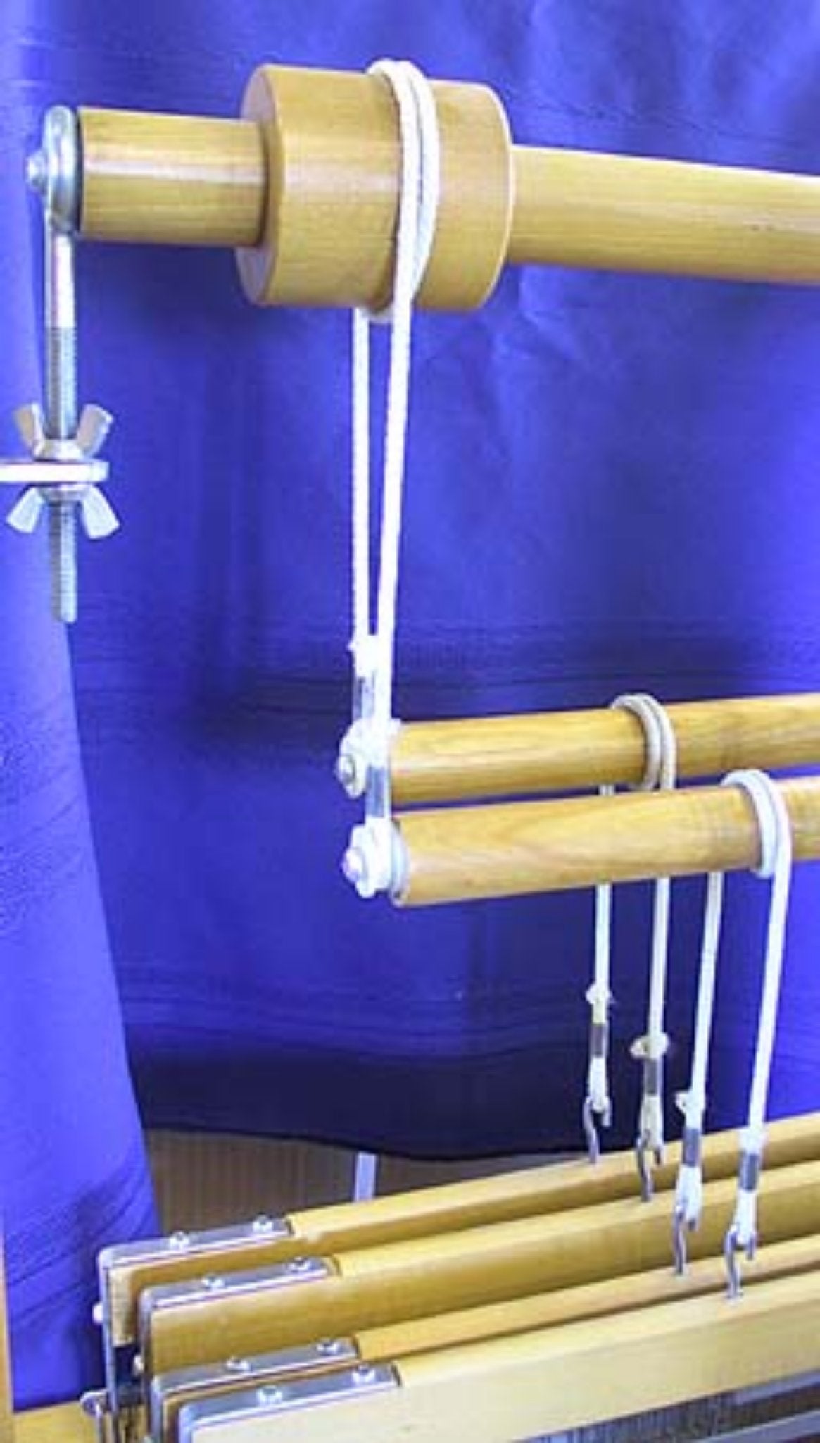 A section of a wooden Leclerc Floor Loom with purple fabric in the background. The loom features several wooden rods and strings with metal clips and bolts, likely used for tension and adjustment, along with a Leclerc White Pulley with screw from Leclerc Looms to aid in the weaving process.