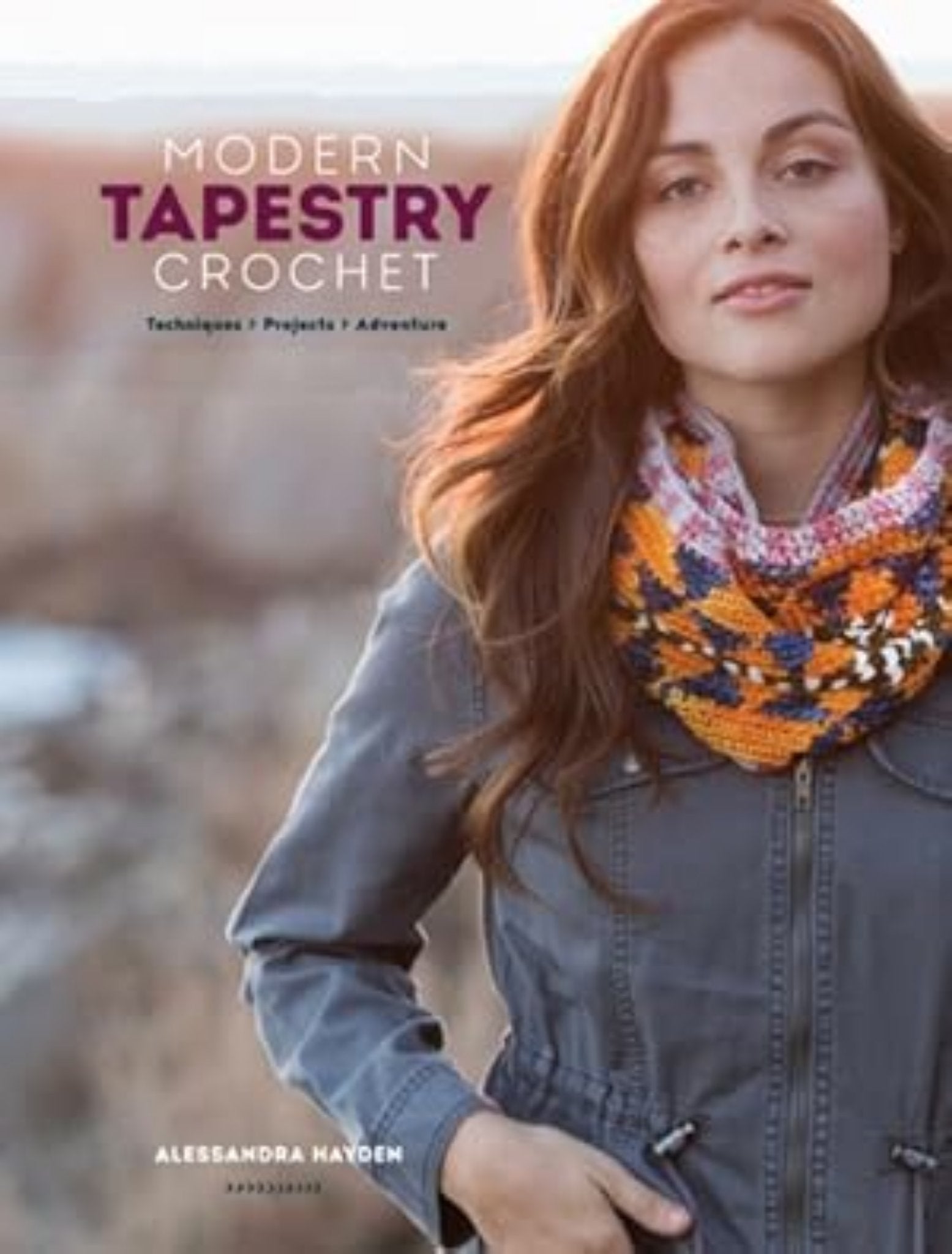 The cover of "Modern Tapestry Crochet" by Alessandra Hayden, published by Ingram Content, showcases a woman with long brown hair modeling a vibrant crocheted scarf that highlights intricate colorwork patterns. She is dressed in a gray jacket and stands against a blurred, natural background.