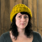 A person with wavy dark hair wears a yellow knit beanie made from Malabrigo Yarn's Rasta merino wool and a grey top, standing in front of a wooden wall, looking directly at the camera.