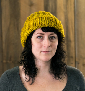 A person with wavy dark hair wears a yellow knit beanie made from Malabrigo Yarn's Rasta merino wool and a grey top, standing in front of a wooden wall, looking directly at the camera.