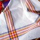 A flat white fabric boasting red, yellow, and purple striped plaid patterns lies seamlessly. A section of a red rope subtly emerges from the left side while a spool of purple thread sits gracefully in the top left corner. Ideal for crafting vibrant napkins or coordinating sets of placemats and napkins using "Best of Handwoven Yarn Series: A Dozen Projects in 8/2 Cotton" by Long Thread Media.