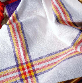 A flat white fabric boasting red, yellow, and purple striped plaid patterns lies seamlessly. A section of a red rope subtly emerges from the left side while a spool of purple thread sits gracefully in the top left corner. Ideal for crafting vibrant napkins or coordinating sets of placemats and napkins using "Best of Handwoven Yarn Series: A Dozen Projects in 8/2 Cotton" by Long Thread Media.