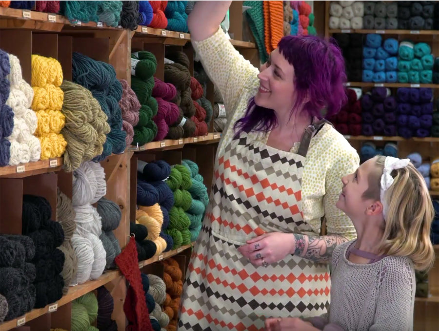 A person with purple hair and a patterned apron reaches for a skein of yarn on a shelf, while a child with a headband watches. They are in a yarn store with shelves filled with various colored yarns.