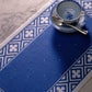 A blue and white patterned table runner is placed on a marble surface. On top of the runner, there is a blue and white teacup on a matching saucer with a silver spoon resting in the cup, cultivating an elegant ambiance similar to that of the projects featured in "Best of Handwoven Yarn Series: A Dozen Projects in 10/2 Pearl Cotton" by Long Thread Media.