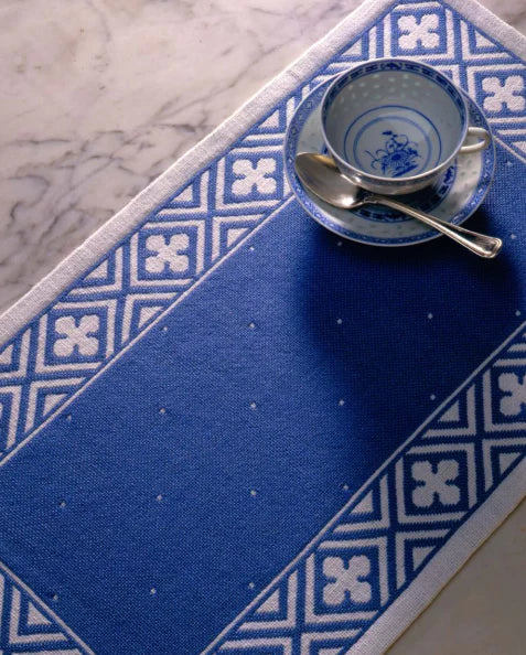 A blue and white patterned table runner is placed on a marble surface. On top of the runner, there is a blue and white teacup on a matching saucer with a silver spoon resting in the cup, cultivating an elegant ambiance similar to that of the projects featured in "Best of Handwoven Yarn Series: A Dozen Projects in 10/2 Pearl Cotton" by Long Thread Media.