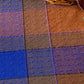 A close-up of the Long Thread Media's Best of Handwoven Yarn Series: A Dozen Projects in 8/2 Cotton, showcasing a multi-colored woven blanket with a checkered pattern in blue, brown, and orange hues. The blanket features a textured design with small raised dots. In the upper right corner, you can see the corner of a wicker basket containing patterned napkins folded inside.