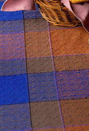 A close-up of the Long Thread Media's Best of Handwoven Yarn Series: A Dozen Projects in 8/2 Cotton, showcasing a multi-colored woven blanket with a checkered pattern in blue, brown, and orange hues. The blanket features a textured design with small raised dots. In the upper right corner, you can see the corner of a wicker basket containing patterned napkins folded inside.