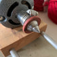 Close-up of a metal tool with a conical tip connected to a gray machine, secured with a clamp. The tool is mounted on a wooden base with metal supports. Red yarn from Marshall Paper Tube Co's Cardboard Spools 4" and a pink circular ring are visible in the background, suggesting efficient yarn management for weavers.