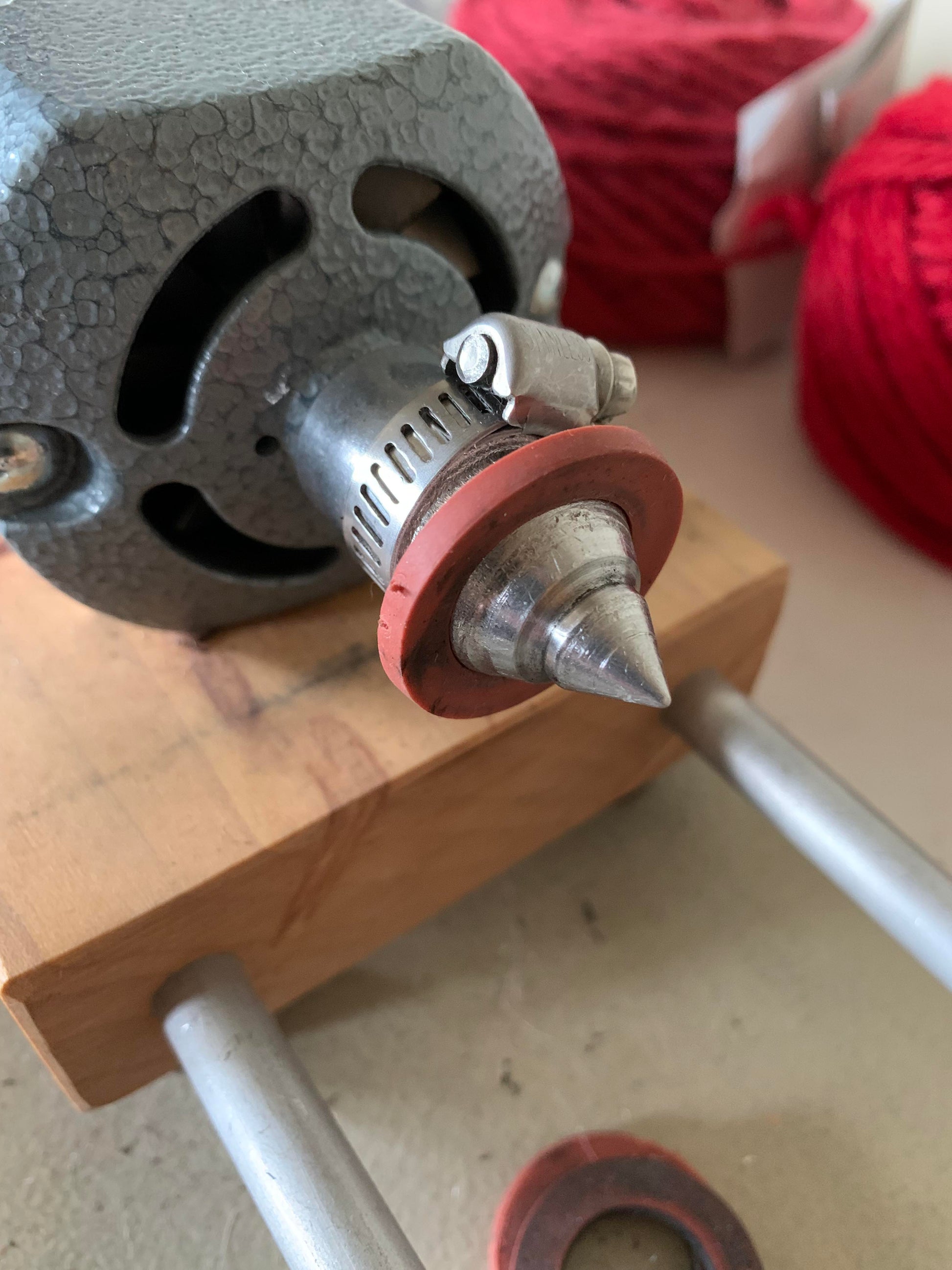 Close-up of a metal tool with a conical tip connected to a gray machine, secured with a clamp. The tool is mounted on a wooden base with metal supports. Red yarn from Marshall Paper Tube Co's Cardboard Spools 4" and a pink circular ring are visible in the background, suggesting efficient yarn management for weavers.