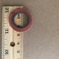 A circular, red rubber gasket lies partially on top of a measuring stick with both metric and imperial units. The gasket's outer diameter spans approximately 12 centimeters (4.7 inches) on the ruler. The surface beneath resembles sturdy Cardboard Spools 4" from Marshall Paper Tube Co, often used for yarn management by weavers.