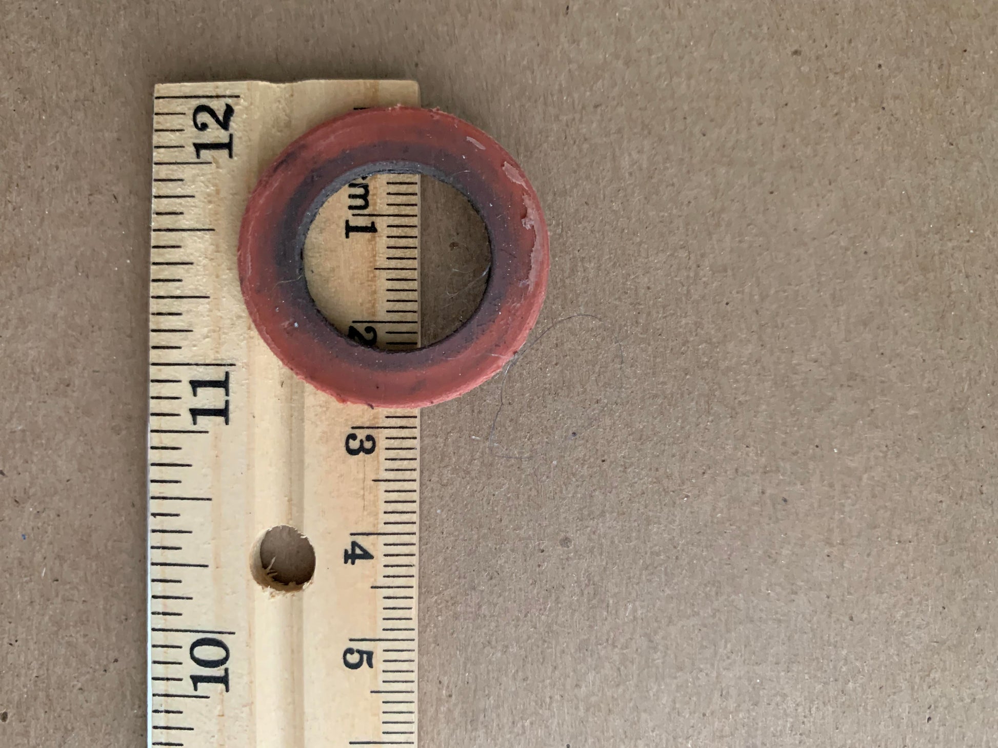 A circular, red rubber gasket lies partially on top of a measuring stick with both metric and imperial units. The gasket's outer diameter spans approximately 12 centimeters (4.7 inches) on the ruler. The surface beneath resembles sturdy Cardboard Spools 4" from Marshall Paper Tube Co, often used for yarn management by weavers.