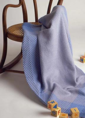 A beautiful light purple blanket crafted from the Best of Handwoven Yarn Series: A Dozen Projects in 8/2 Cotton by Long Thread Media is draped over a wooden chair with a wicker seat. Wooden alphabet blocks with letters 'W', 'F', and 'I' are scattered on the floor beside the chair, alongside neatly folded patterned napkins.