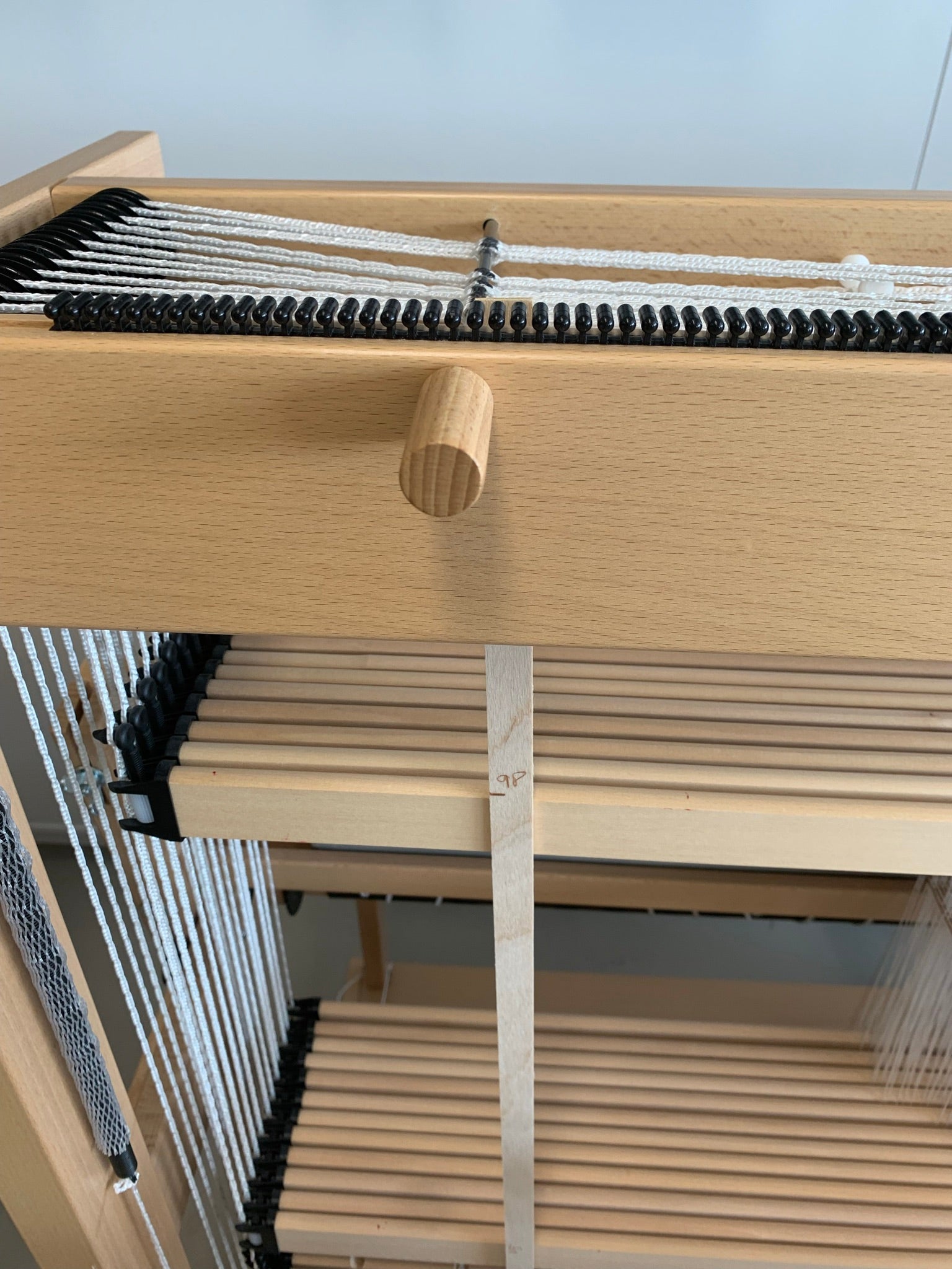 A close-up of a Louët Measuring Rod Hollandia by Louët Inc. showcases a section with black and white threads stretched across it. A wooden peg is positioned in the middle, with a series of wooden slats below the threads providing perfect storage, all within a light wood structure.