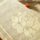 A close-up view of the beige crocheted tablecloth, made from the "Best of Handwoven Yarn Series: A Dozen Projects in 10/2 Pearl Cotton (Printed Version)" by Long Thread Media, showcases its subtle floral design. Two colorful Christmas ornaments on the upper part add a festive touch, while the texture and intricate patterns of the crochet work are clearly visible.