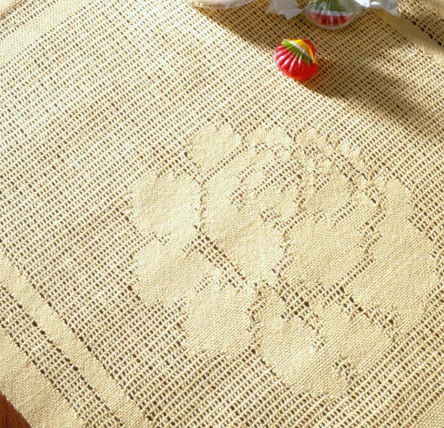 A close-up view of the beige crocheted tablecloth, made from the "Best of Handwoven Yarn Series: A Dozen Projects in 10/2 Pearl Cotton (Printed Version)" by Long Thread Media, showcases its subtle floral design. Two colorful Christmas ornaments on the upper part add a festive touch, while the texture and intricate patterns of the crochet work are clearly visible.