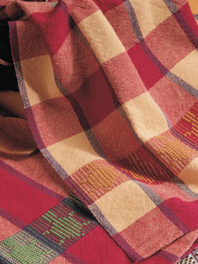 A close-up image of Best of Handwoven Yarn Series: A Dozen Projects in 8/2 Cotton (Printed Version) by Long Thread Media, showcasing a colorful checkered pattern reminiscent of patterned napkins. The fabric displays large squares in shades of red, beige, green, and blue, with intricate detailing and textures visible. The material appears to be draped casually.