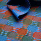 A close-up of the Best of Handwoven Yarn Series: A Dozen Projects in 10/2 Pearl Cotton from Long Thread Media showcases a vibrant textile with an interlocking pattern of green, blue, and orange circles and rectangles. One corner of the fabric is folded over, revealing part of a silver bowl at the top of the image.