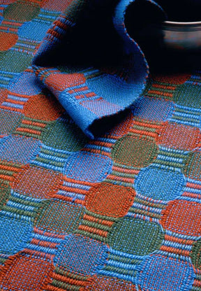 A close-up of the Best of Handwoven Yarn Series: A Dozen Projects in 10/2 Pearl Cotton from Long Thread Media showcases a vibrant textile with an interlocking pattern of green, blue, and orange circles and rectangles. One corner of the fabric is folded over, revealing part of a silver bowl at the top of the image.